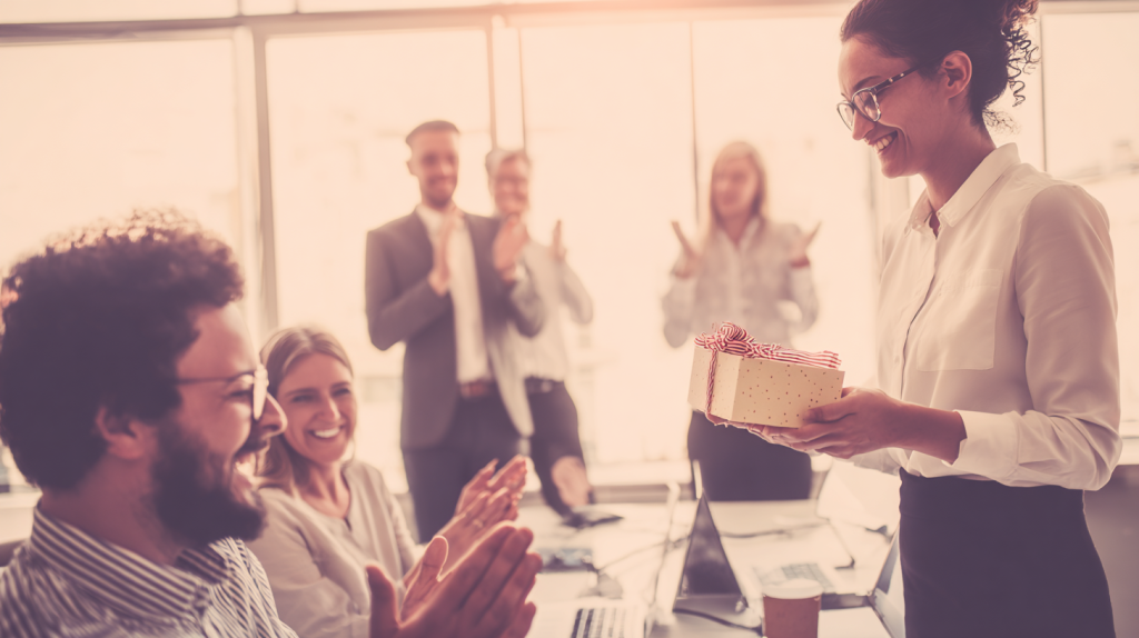 A manager hands a wrapped gift to an employee in a modern office while coworkers clap and smile in the background, illustrating employee recognition and appreciation as a workplace celebration idea.
