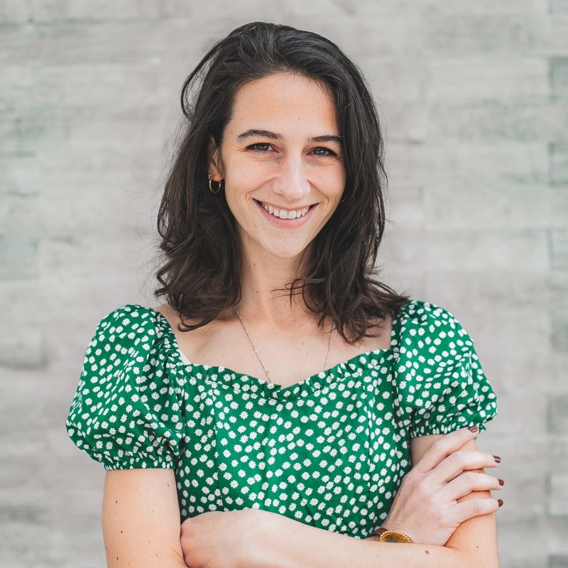 Portrait of Abigail Caldwell, a People Operations and HR professional, smiling confidently with arms crossed. She has shoulder-length dark hair, is wearing a green patterned dress with puff sleeves, and is standing against a light gray textured wall.