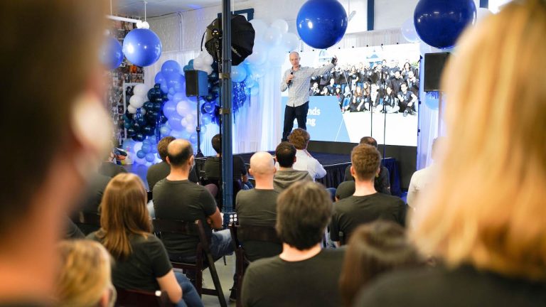 CEO presenting at a company all-hands meeting in front of team members and a balloon-decorated stage
